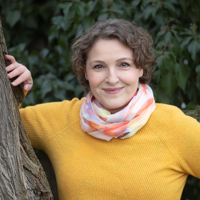 Bild der Autorin Nina Blazon, die an einen Baum gelehnt ist mit gelbem Pulli und Schal